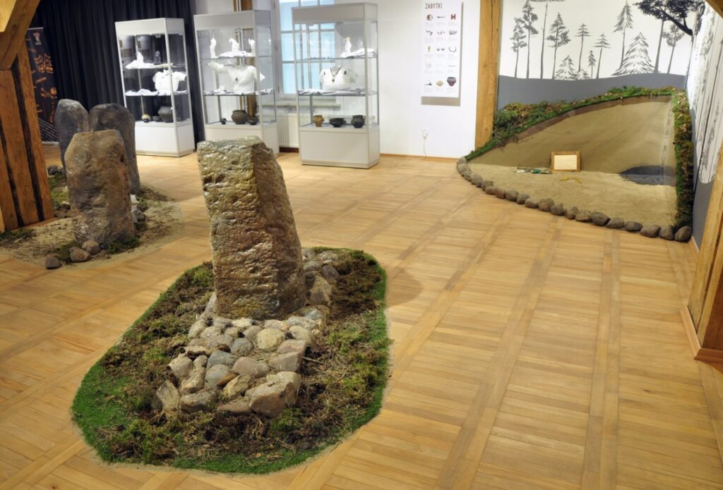 Widok ekspozycji łączącej kamienne stele z przeszklonymi gablotami i rozbudowaną scenografią krajobrazową; na pierwszym planie znajduje się kamienny obiekt otoczony żwirem, kamieniami i fragmentami zieleni; w tle widoczne są gabloty z zabytkami oraz pagórkowata forma przedstawiająca element dawnego krajobrazu (kliknięcie powiększy zdjęcie).