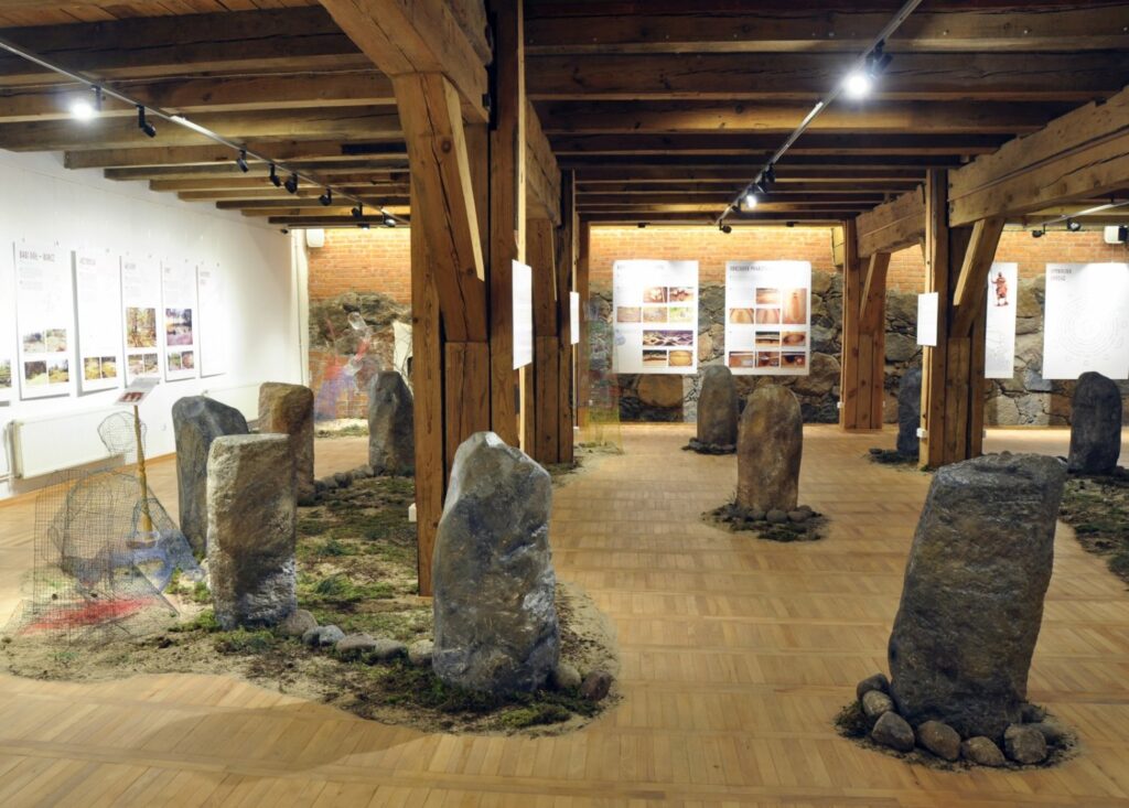 Widok sali wystawowej zaaranżowanej wokół kamiennych głazów ustawionych w niewielkich, naturalistycznych kompozycjach; kamienne elementy otaczają fragmenty zieleni, ziemi i żwiru, tworząc nawiązanie do dawnego krajobrazu i archeologicznych miejsc kultu; w tle widoczne są plansze informacyjne, fotografie oraz inne elementy ekspozycji; drewniana konstrukcja (kliknięcie powiększy zdjęcie).