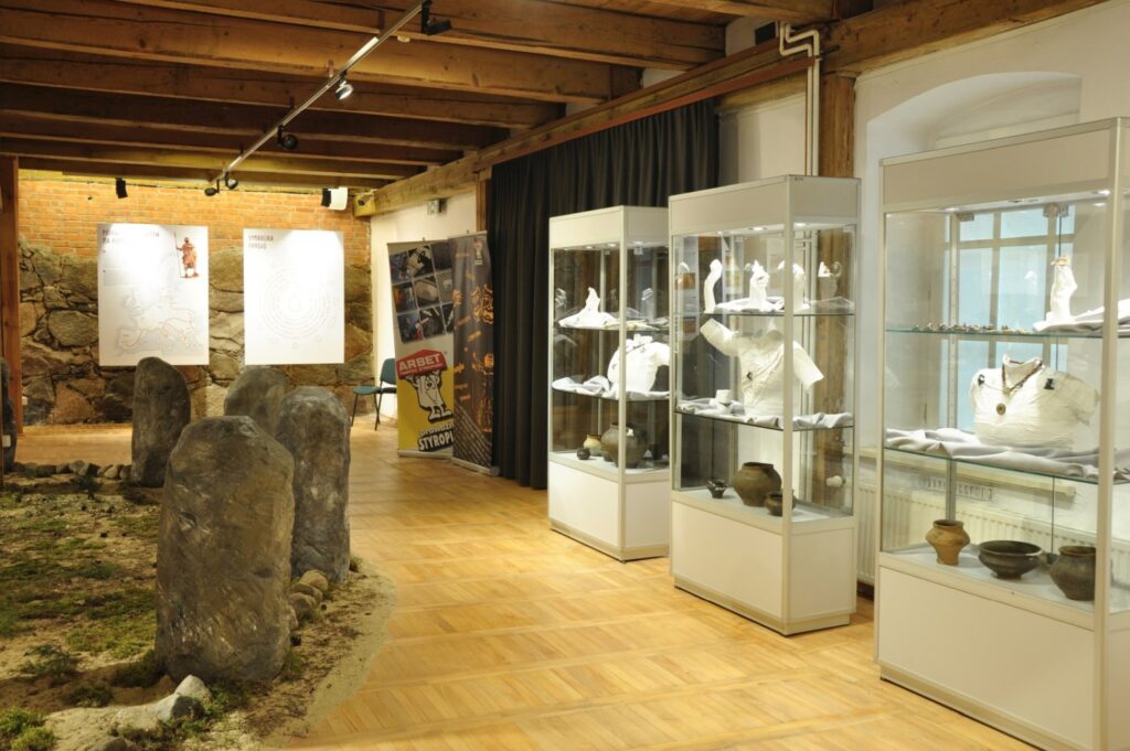 Po prawej stronie znajdują się przeszklone gabloty z różnorodnymi przedmiotami, w tym naczyniami i obiektami archeologicznymi; w centralnej części sali ustawiono kamienne stele otoczone żwirem i roślinnością; na ścianach prezentowane są materiały ilustracyjne i informacyjne uzupełniające wystawę (kliknięcie powiększy zdjęcie).