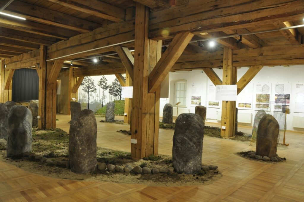 Fragment wystawy z kamiennymi stelami rozmieszczonymi w kilku grupach na powierzchni imitującej naturalne podłoże; pomiędzy kamieniami znajdują się fragmenty trawy, ziemi i żwiru, dzięki czemu ekspozycja przypomina stanowisko archeologiczne w plenerze; w tle widoczne są gabloty, plansze oraz ilustracje związane z prezentowaną tematyką (kliknięcie powiększy zdjęcie).