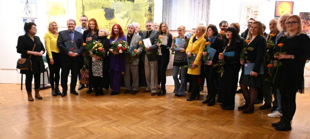Fotografia grupowa uczestników wernisażu; na tle prezentowanych prac pozują artyści, organizatorzy oraz goście wydarzenia, część osób trzyma katalogi wystawy, kwiaty i pojedyncze róże; zdjęcie dokumentuje wspólne spotkanie i uroczyste otwarcie wystawy w przestrzeni muzealnej (kliknięcie powiększy zdjęcie).