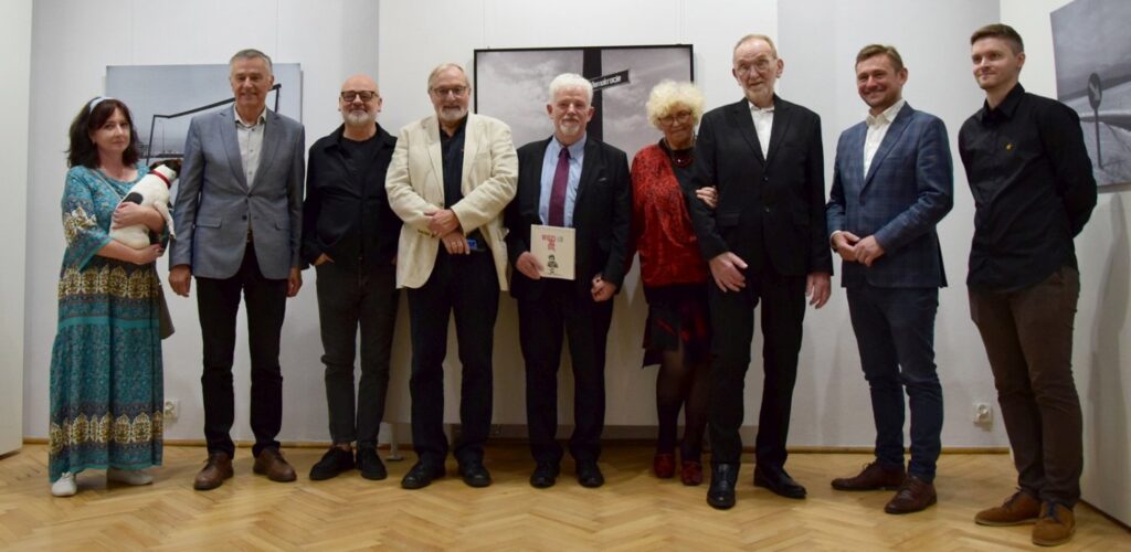 Grupa dziewięciu osób pozuje na tle jasnej ściany do wspólnego, pamiątkowego zdjęcia; po lewej stoi kobieta, która trzyma na rękach małego psa (kliknięcie powiększy zdjęcie).