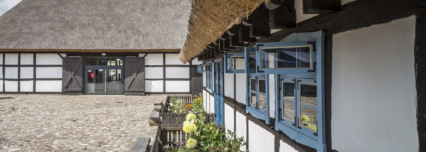 Dwa budynki stylizowane na architekturę szachulcową. Po prawej stronie widoczne okna, a przed budynkiem kolorowe kwiaty (kliknięcie spowoduje przejście do zakładki z opisem Zagrody Jamneńskiej).