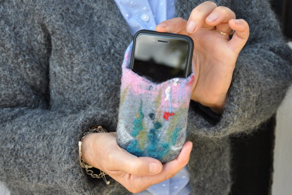 Kobieta trzyma w dłoni etui na telefon wykonane z filcu. W etui tym umieszczony jest telefon (kliknięcie spowoduje powiększenie zdjęcia)
