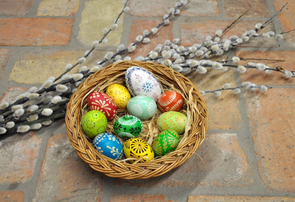 W koszyczku znajdują się kolorowe wielkanocne pisanki. Koszyczek położony jest na ceglanej podłodze, a obok niego leżą gałązki wierzby z baziami (kliknięcie spowoduje powiększenie zdjęcia)