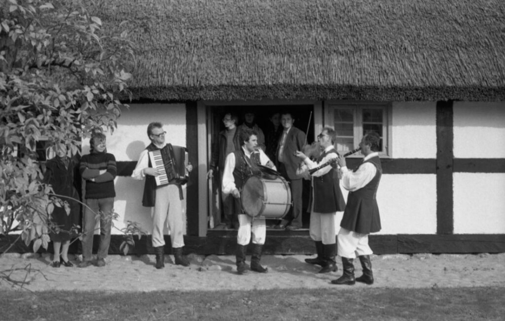 Zespół ludowy grający muzykę przed tradycyjnym budynkiem skansenowym podczas uroczystości otwarcia, w tle zgromadzeni uczestnicy (kliknięcie powiększy zdjęcie)