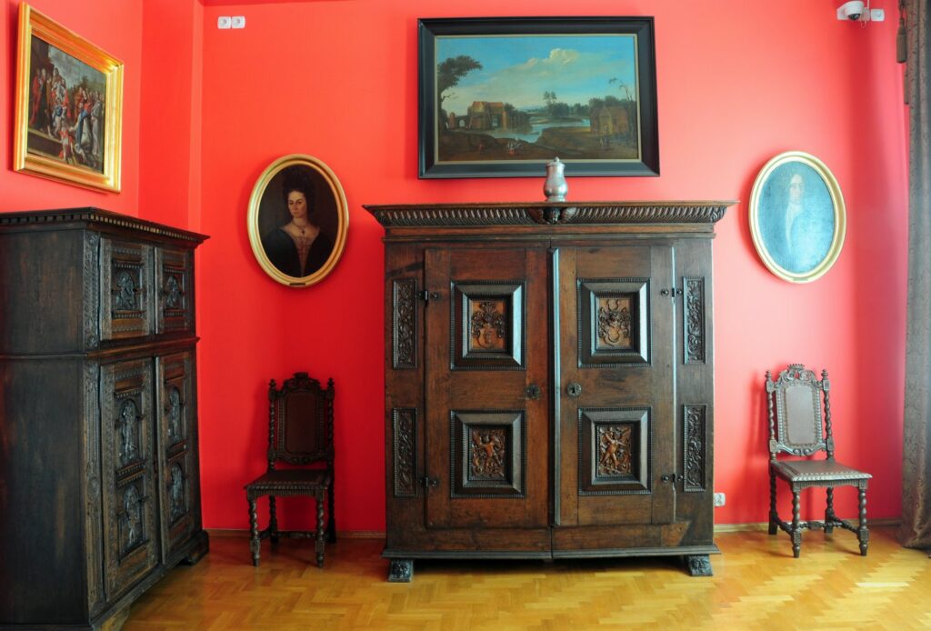 Wnętrze w stylu barokowym z dużą, rzeźbioną drewnianą szafą, obrazami portretowymi na czerwonej ścianie oraz zabytkowym drewnianym krzesłem (kliknięcie powiększy zdjęcie)