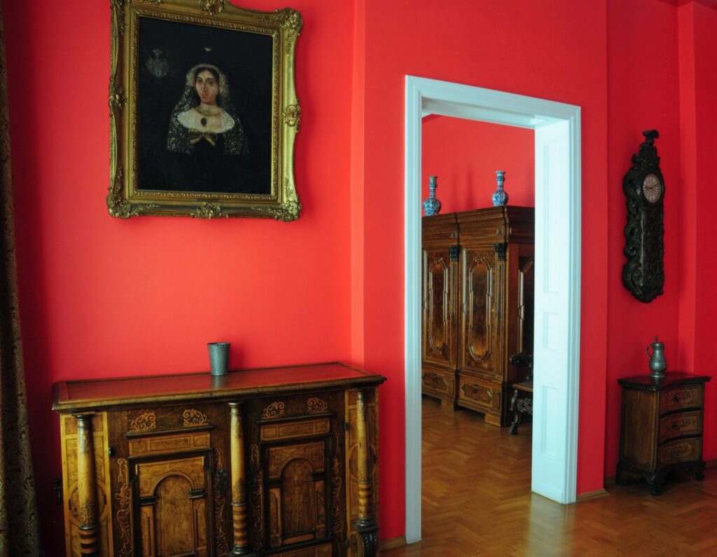 Zabytkowa komoda i portret na czerwonej ścianie, z widokiem przez drzwi na drugi pokój urządzony barokowymi meblami (kliknięcie powiększy zdjęcie)