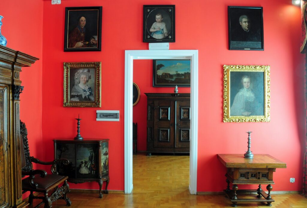 Ściana z portretami i widok przez otwarte drzwi na drugi pokój z drewnianą szafą (kliknięcie powiększy zdjęcie)