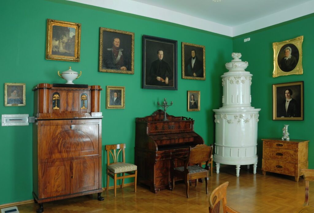 Wnętrze muzealne z zielonymi ścianami: pianino, biała kaflowa kolumna, drewniane meble oraz portrety w ramach zawieszone na ścianie (kliknięcie powiększy zdjęcie)