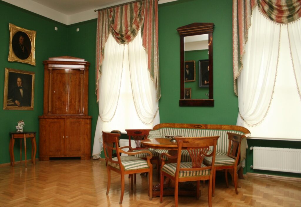 Historyczny salon z zielonymi ścianami: okrągły stół, krzesła, kanapa, wysokie okno z dekoracyjnymi zasłonami oraz lustro na ścianie (kliknięcie powiększy zdjęcie)