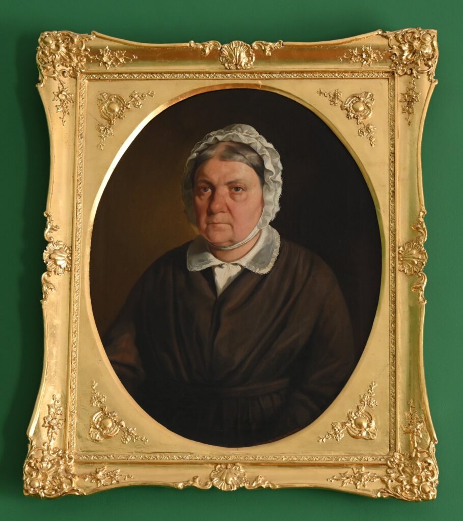 Portret starszej kobiety w ciemnym stroju, ujęty w ozdobną złoconą ramę, zawieszony na zielonej ścianie (kliknięcie powiększy zdjęcie)