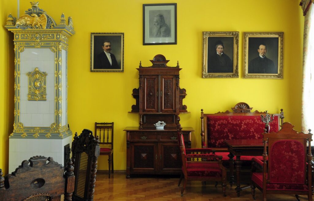 Sala wystawowa z żółtymi ścianami: drewniane meble, kredens, stół, krzesła oraz portrety w ramach (kliknięcie powiększy zdjęcie)