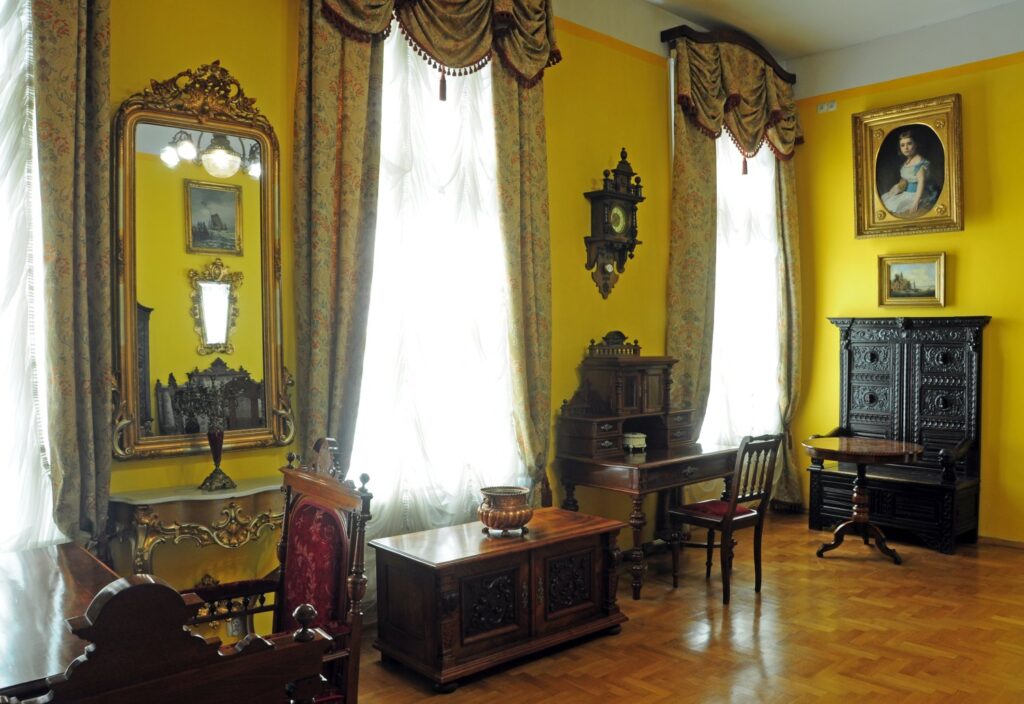 Sala wystawowa z żółtymi ścianami: lustro w złoconej ramie, drewniane biurko, krzesła, okno z zasłonami oraz obrazy na ścianach (kliknięcie powiększy zdjęcie)