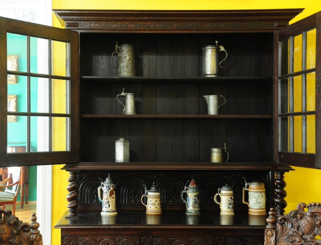 Otwarta drewniana witryna z półkami, na których stoją ceramiczne i metalowe naczynia, na tle żółtej ściany (kliknięcie powiększy zdjęcie)
