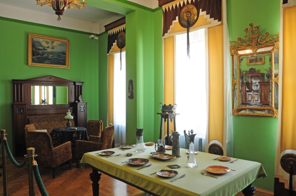 Historyczna jadalnia z zielonymi ścianami: stół nakryty obrusem i zastawą, tapicerowane fotele, obrazy na ścianach oraz wysokie okna (kliknięcie powiększy zdjęcie)