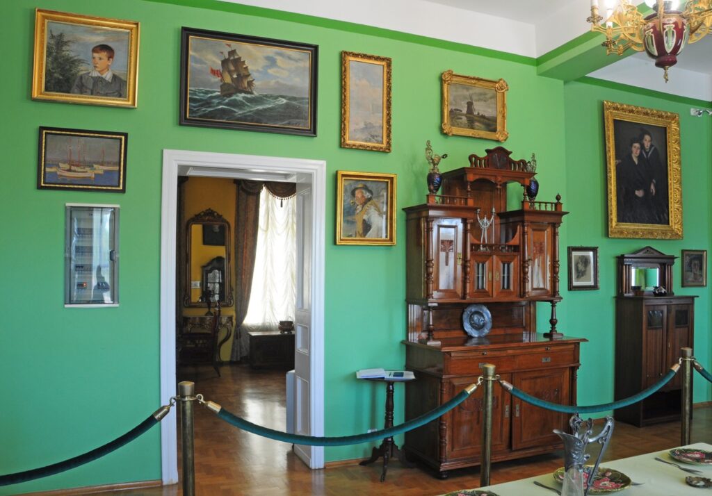 Muzealne wnętrze z zielonymi ścianami: drewniany kredens, liczne obrazy i ryciny w ramach, z boku przejście do kolejnego pomieszczenia (kliknięcie powiększy zdjęcie)