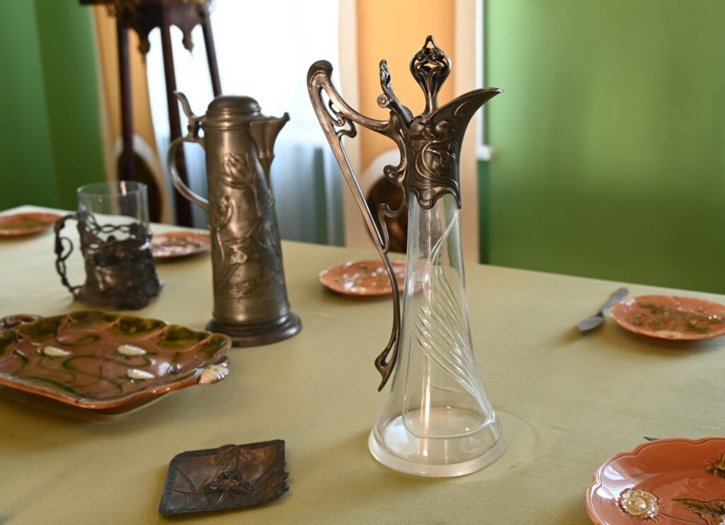 Zbliżenie na stół z zastawą: ceramiczne talerze, metalowe naczynia i drobne przedmioty użytkowe na jasnym obrusie (kliknięcie powiększy zdjęcie)