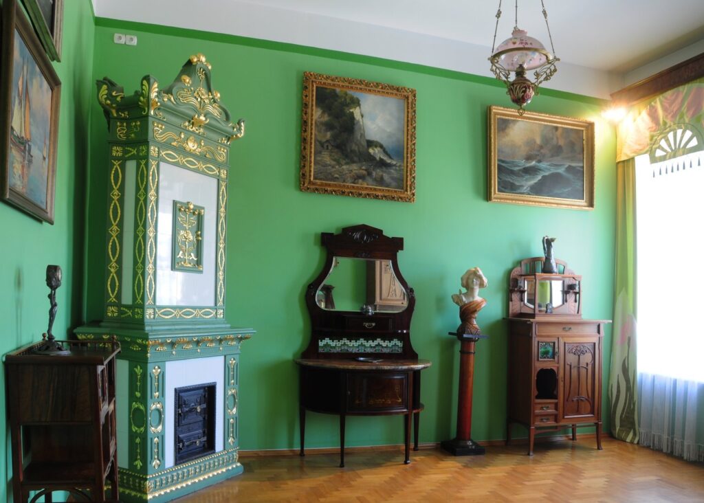 Wnętrze pokoju z zielonymi ścianami: dekoracyjny piec kaflowy, lustro, drewniane meble, rzeźba oraz obrazy marynistyczne (kliknięcie powiększy zdjęcie)