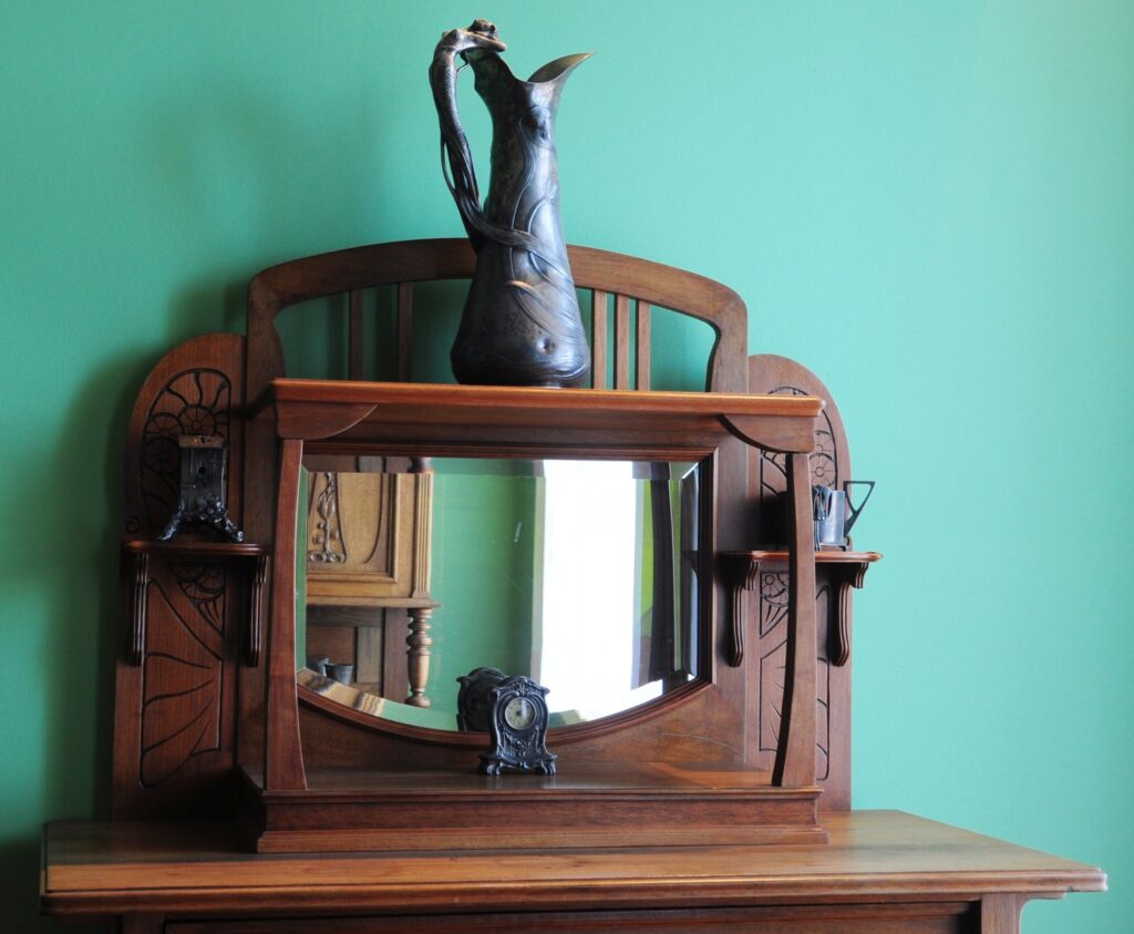 Drewniana toaletka z lustrem i rzeźbioną dekoracją, na której stoi ciemna figura ptaka, na tle zielonej ściany (kliknięcie powiększy zdjęcie)