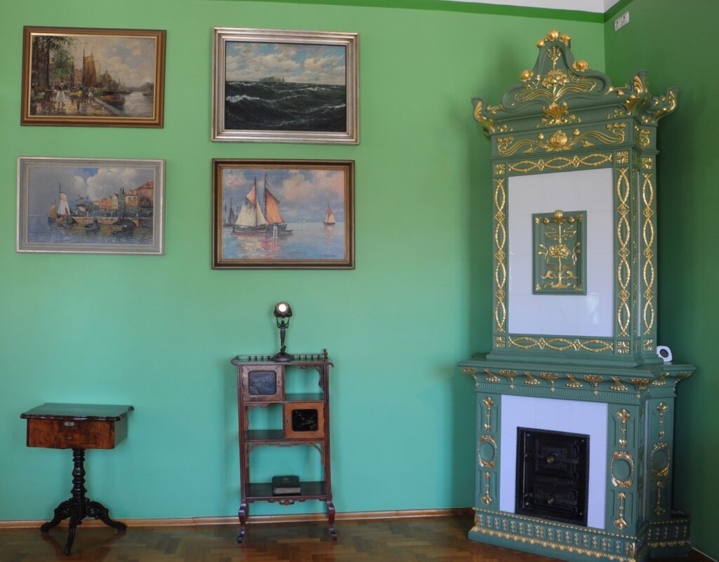 Ściana pokoju z zielonymi ścianami: kaflowy kominek, kilka obrazów przedstawiających statki oraz niewielkie stoliki i meble (kliknięcie powiększy zdjęcie)