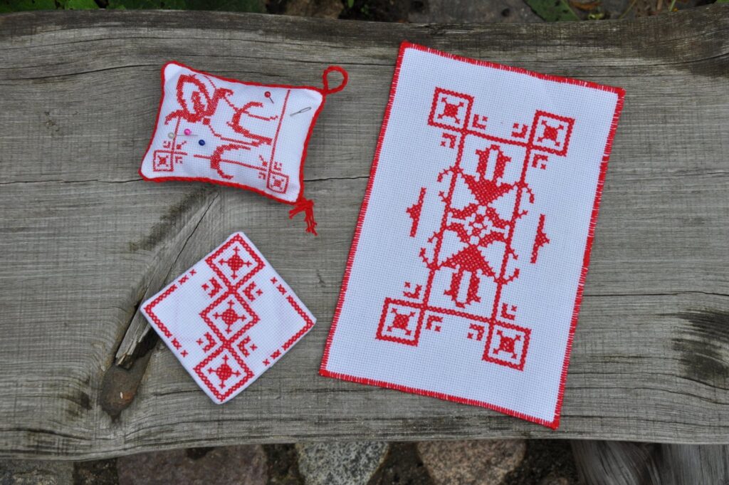 Na drewnianej desce leżą trzy wzory haftu krzyżykowego w kolorze czerwonym, na białym tle (kliknięcie spowoduje powiększenie zdjęcia)
