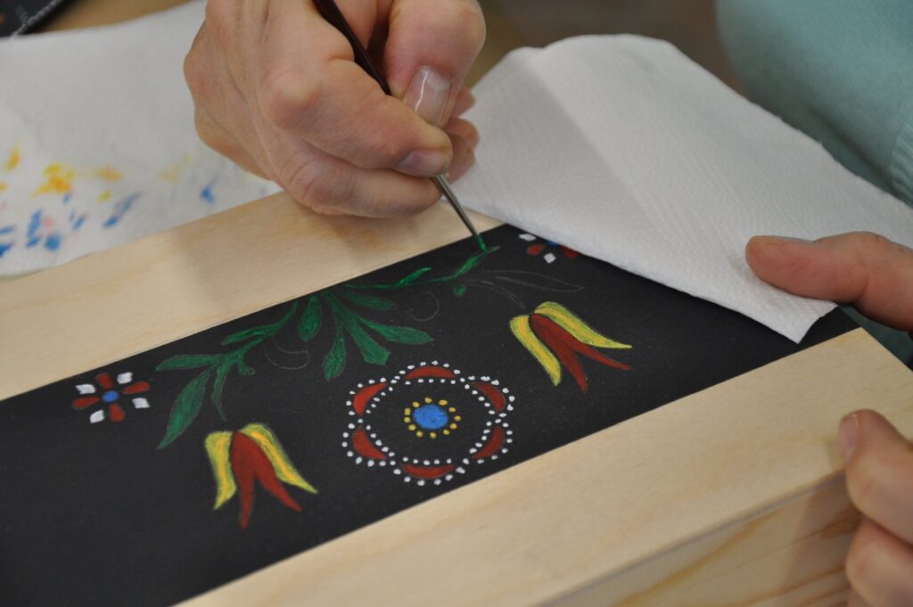 Kobieta trzyma w dłoni narzędzie, którym maluje polichromię na desce, na czarnym tle (kliknięcie spowoduje powiększenie zdjęcia)