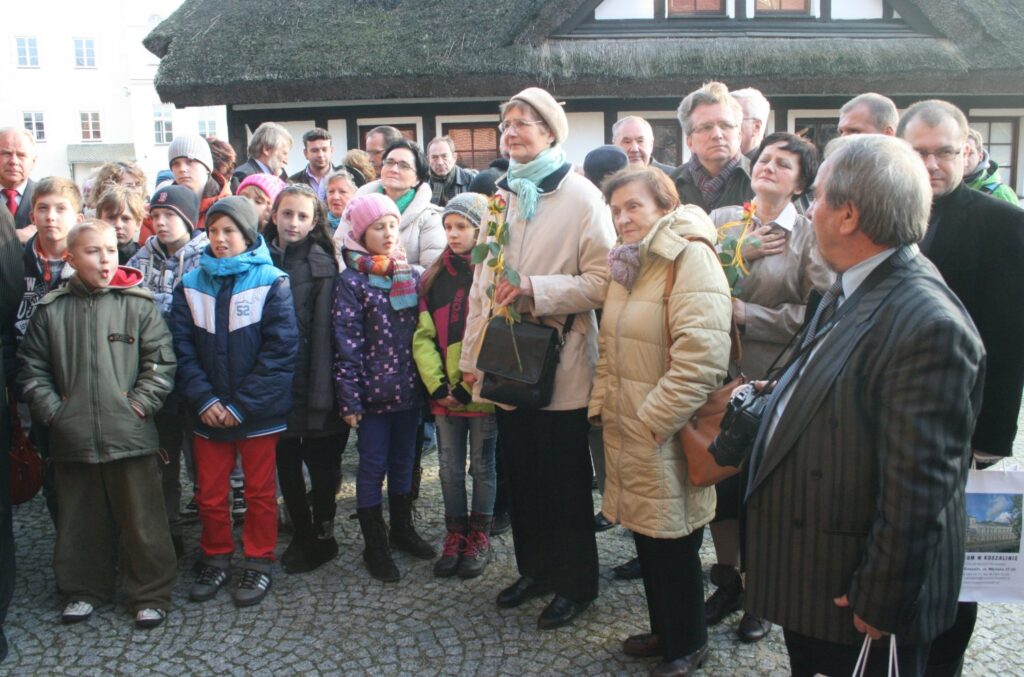 Grupa dorosłych i dzieci słuchających wystąpienia podczas otwarcia wystawy na terenie muzeum (kliknięcie powiększy zdjęcie)
