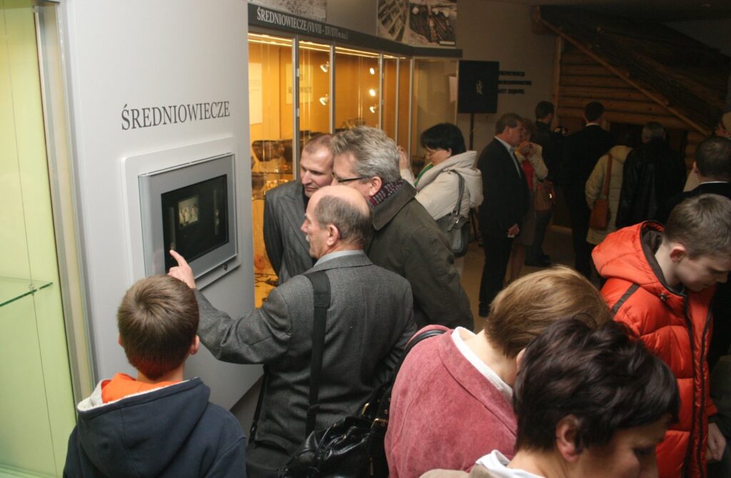 Grupa zwiedzających skupiona wokół interaktywnego stanowiska multimedialnego na wystawie (kliknięcie powiększy zdjęcie)