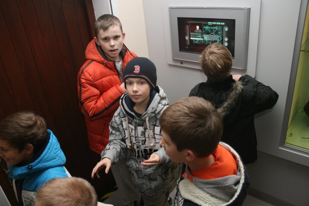 Dzieci stojące przy ekranie multimedialnym prezentującym treści wystawy (kliknięcie powiększy zdjęcie)