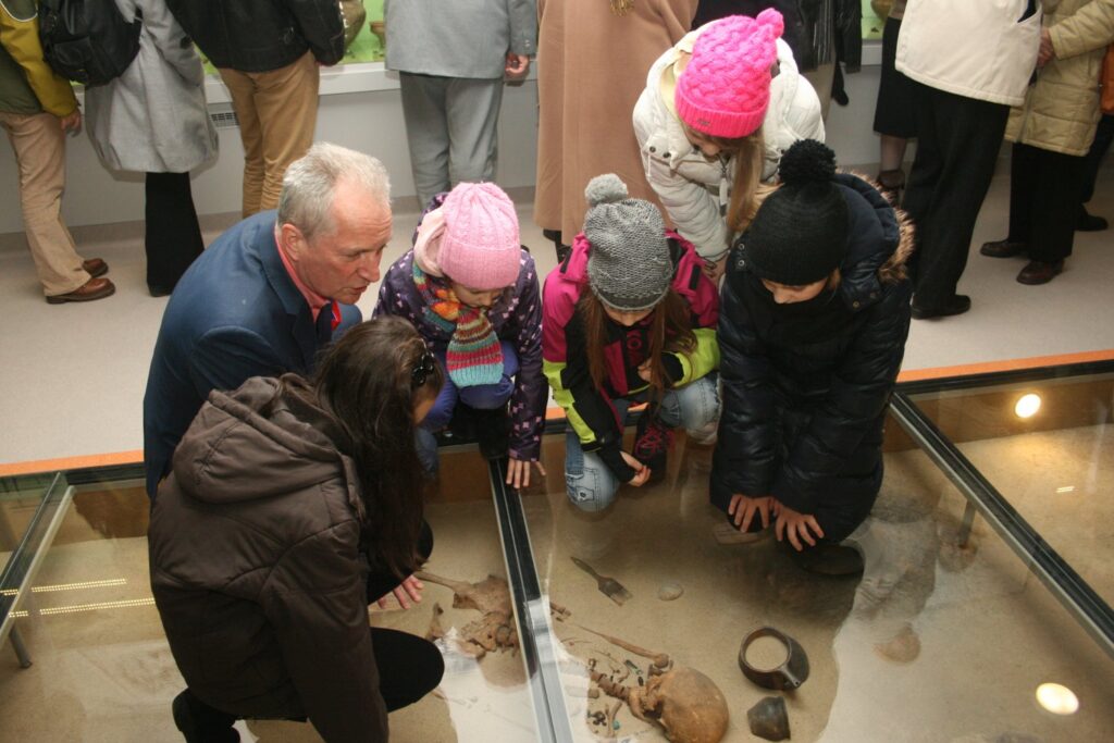 Zwiedzający pochylający się nad przeszkloną gablotą podłogową z zabytkami, wśród nich dzieci i dorośli (kliknięcie powiększy zdjęcie)