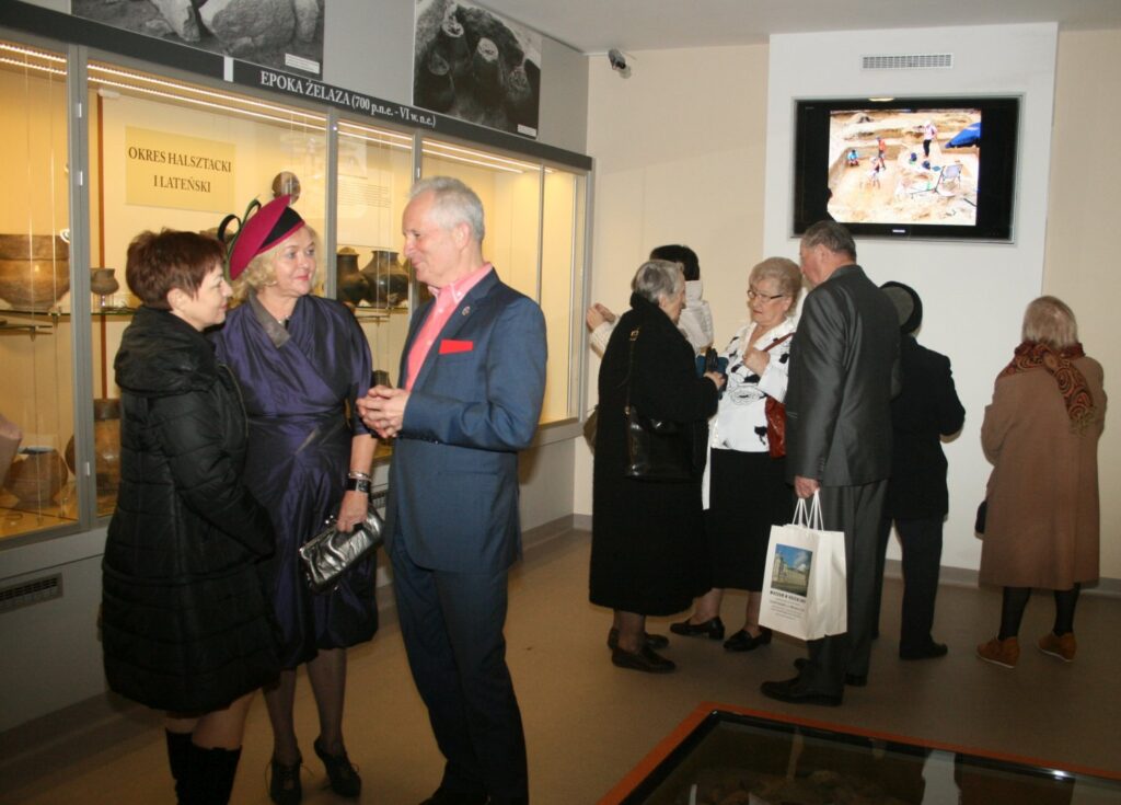 Rozmowa gości w sali wystawowej podczas otwarcia ekspozycji, w tle gabloty z zabytkami (kliknięcie powiększy zdjęcie)