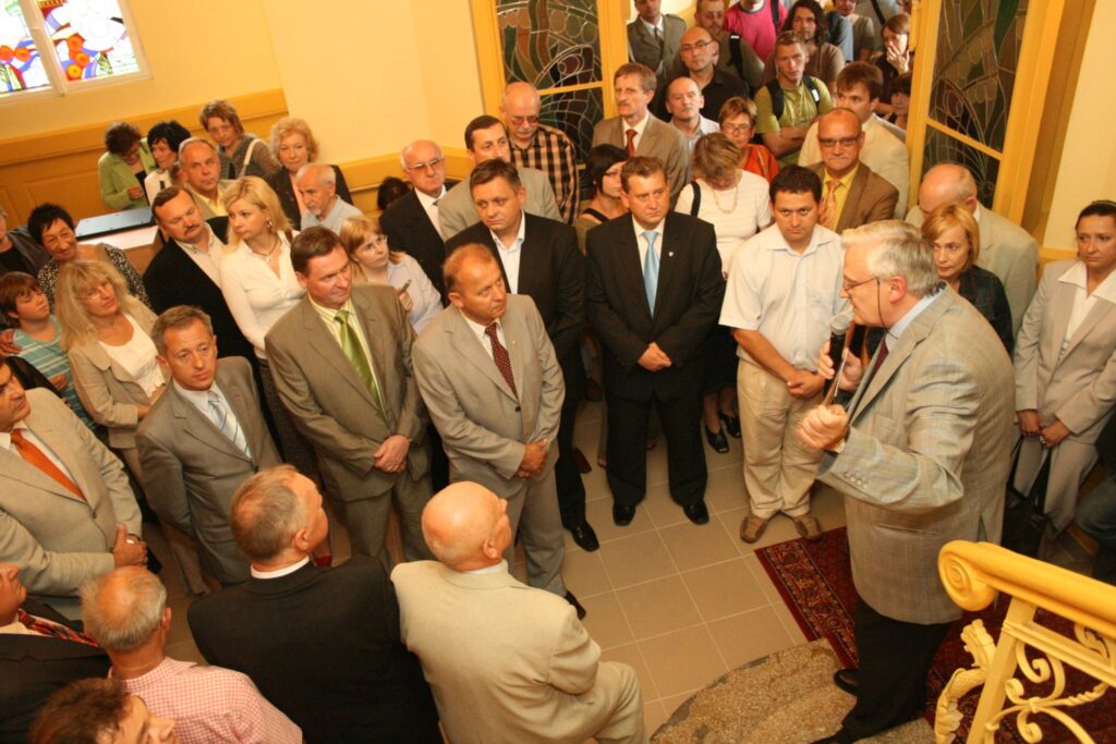 Grupa osób zgromadzona na korytarzu podczas wydarzenia, stojąca i słuchająca przemawiającego mężczyzny (kliknięcie powiększy zdjęcie)