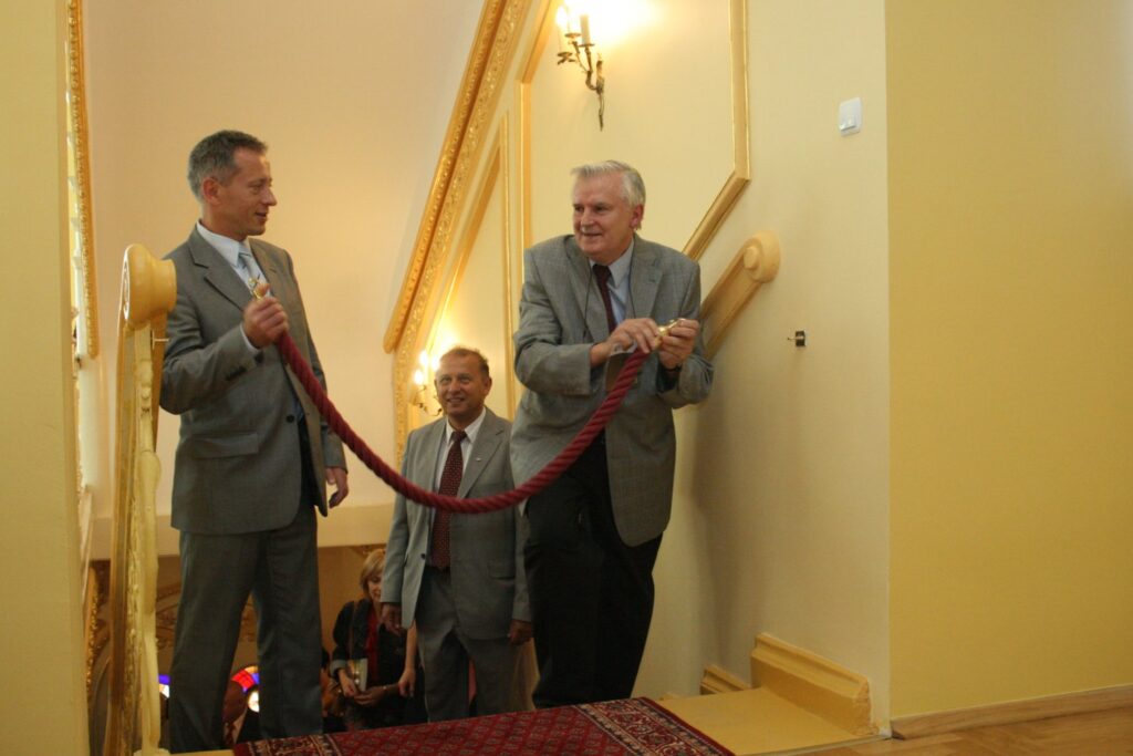 Dwóch mężczyzn trzymających czerwoną wstęgę podczas uroczystości otwarcia na klatce schodowej (kliknięcie powiększy zdjęcie)