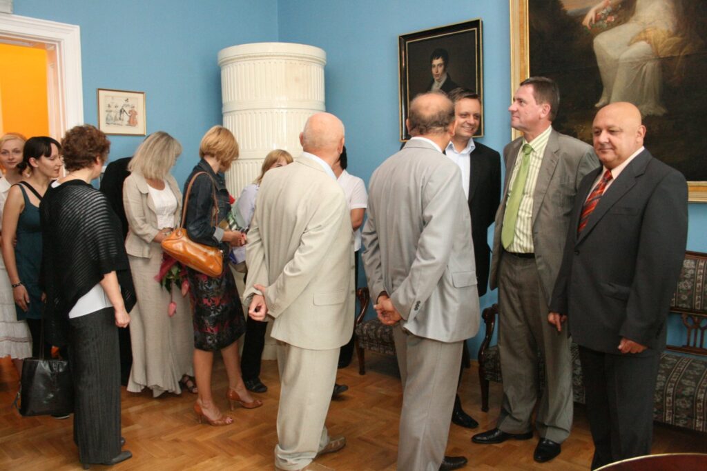 Grupa osób stojąca w muzealnej sali z niebieskimi ścianami i portretami, rozmawiająca podczas wydarzenia (kliknięcie powiększy zdjęcie)