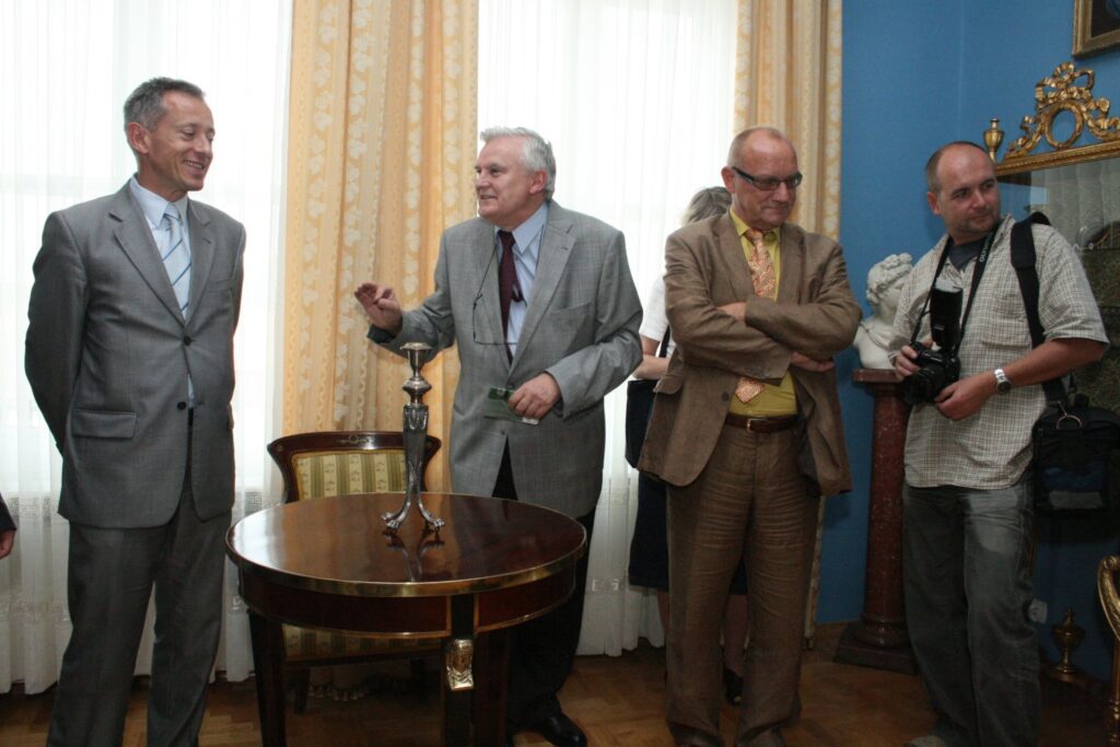 Kilka osób stojących w muzealnym wnętrzu przy oknie, rozmawiających obok okrągłego stołu i zabytkowych mebli (kliknięcie powiększy zdjęcie)