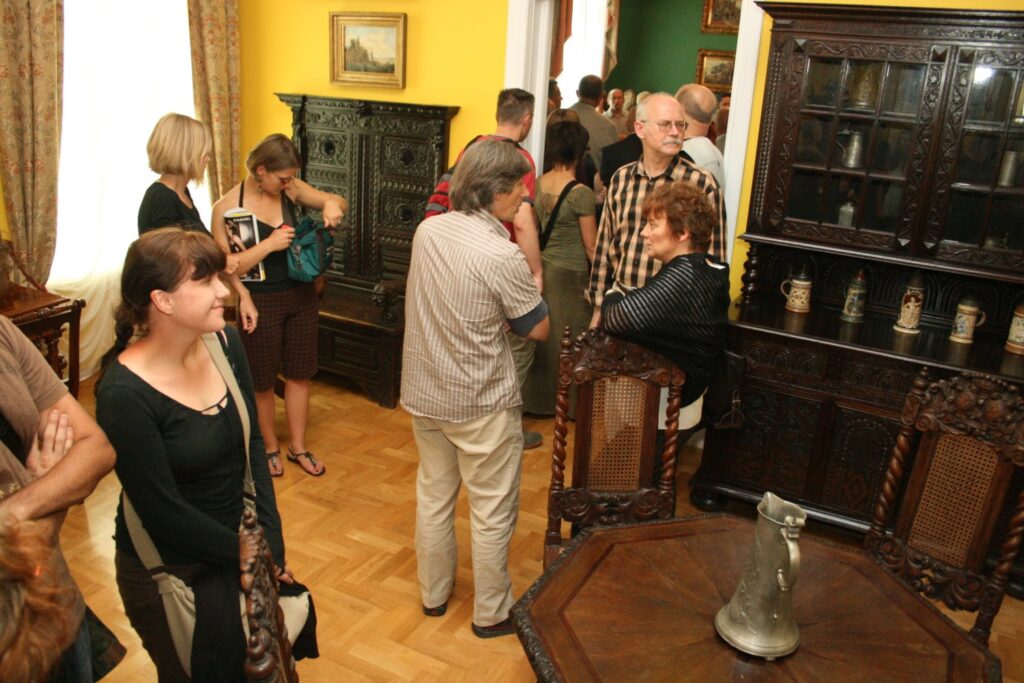 Osoby zwiedzające muzealną salę z ciemnymi meblami i żółtą ścianą, stojące i oglądające eksponaty (kliknięcie powiększy zdjęcie)