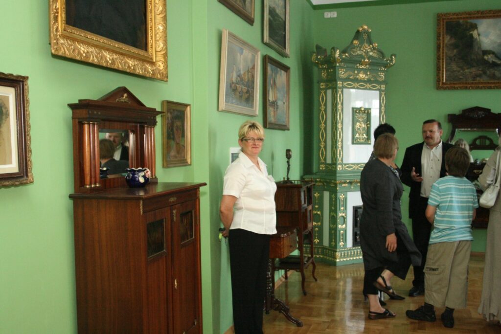 Osoby rozmawiające w muzealnym wnętrzu o zielonych ścianach z licznymi obrazami i zabytkowym piecem kaflowym (kliknięcie powiększy zdjęcie)