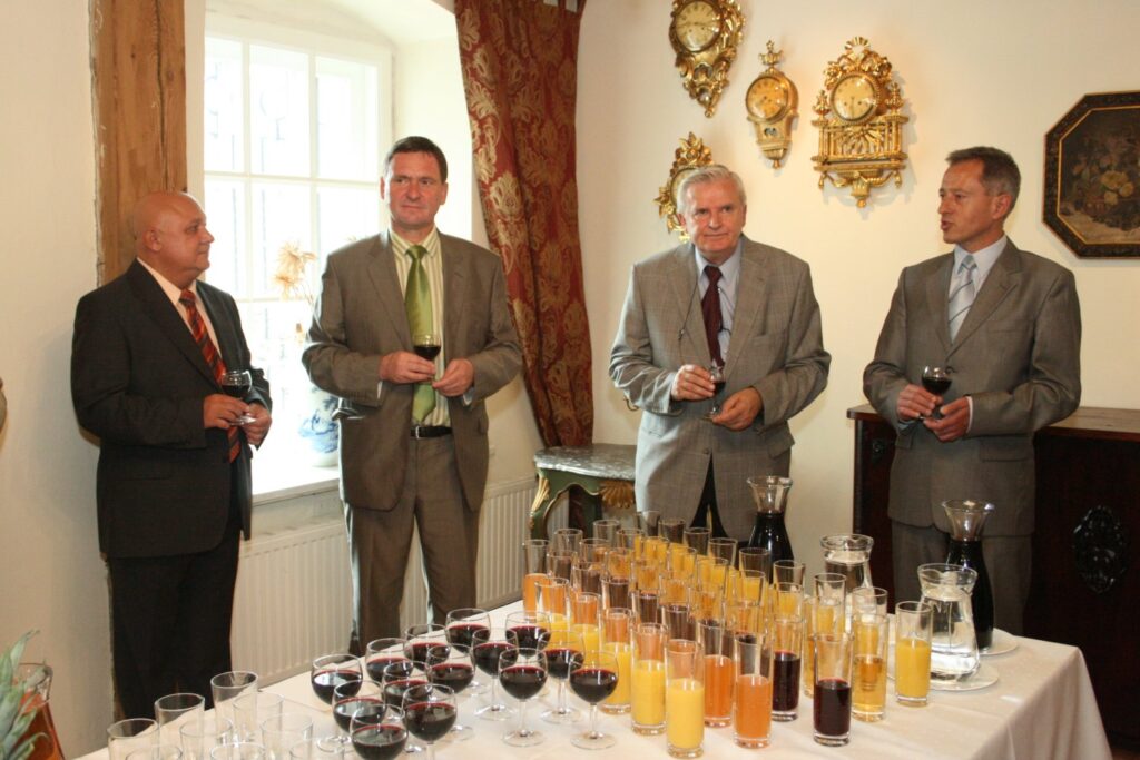 Czterech mężczyzn stojących przy stole z napojami w muzealnej sali z białą ścianą i dekoracjami ściennymi (kliknięcie powiększy zdjęcie)