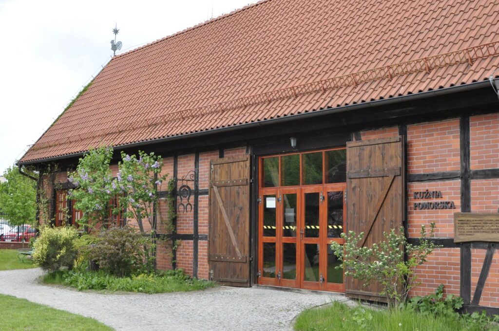 Budynek kuźni w skansenie – ceglana ściana z drewnianą konstrukcją szkieletową, dwuspadowy dach z dachówki, wejście z drewnianymi drzwiami (kliknięcie powiększy zdjęcie)