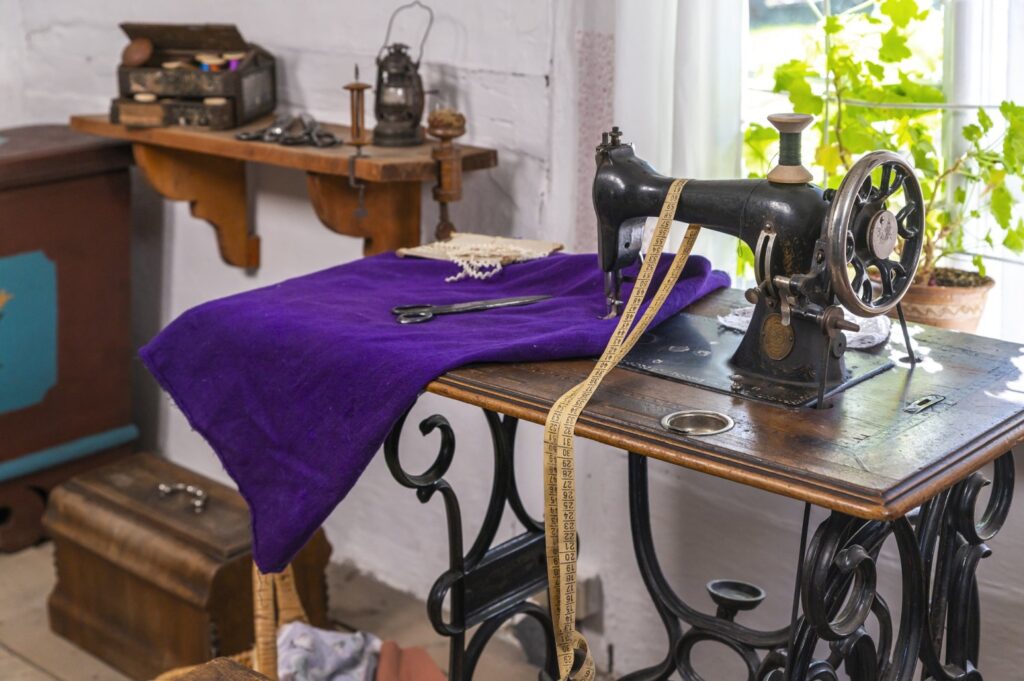 Maszyna do szycia z początku XX wieku ustawiona na stole, z materiałami krawieckimi jako element wyposażenia chałupy (kliknięcie powiększy zdjęcie).
