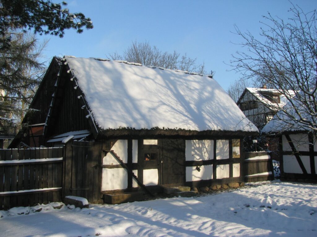 Budynek warsztatu szewskiego w skansenie, chałupa z dwuspadowym dachem, otoczona zimowym krajobrazem (kliknięcie powiększy zdjęcie).