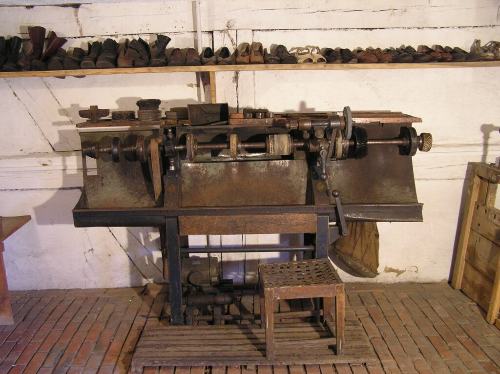 Maszyna szewska do obróbki obuwia ustawiona we wnętrzu warsztatu, z widocznym mechanizmem i półką na buty w tle (kliknięcie powiększy zdjęcie).