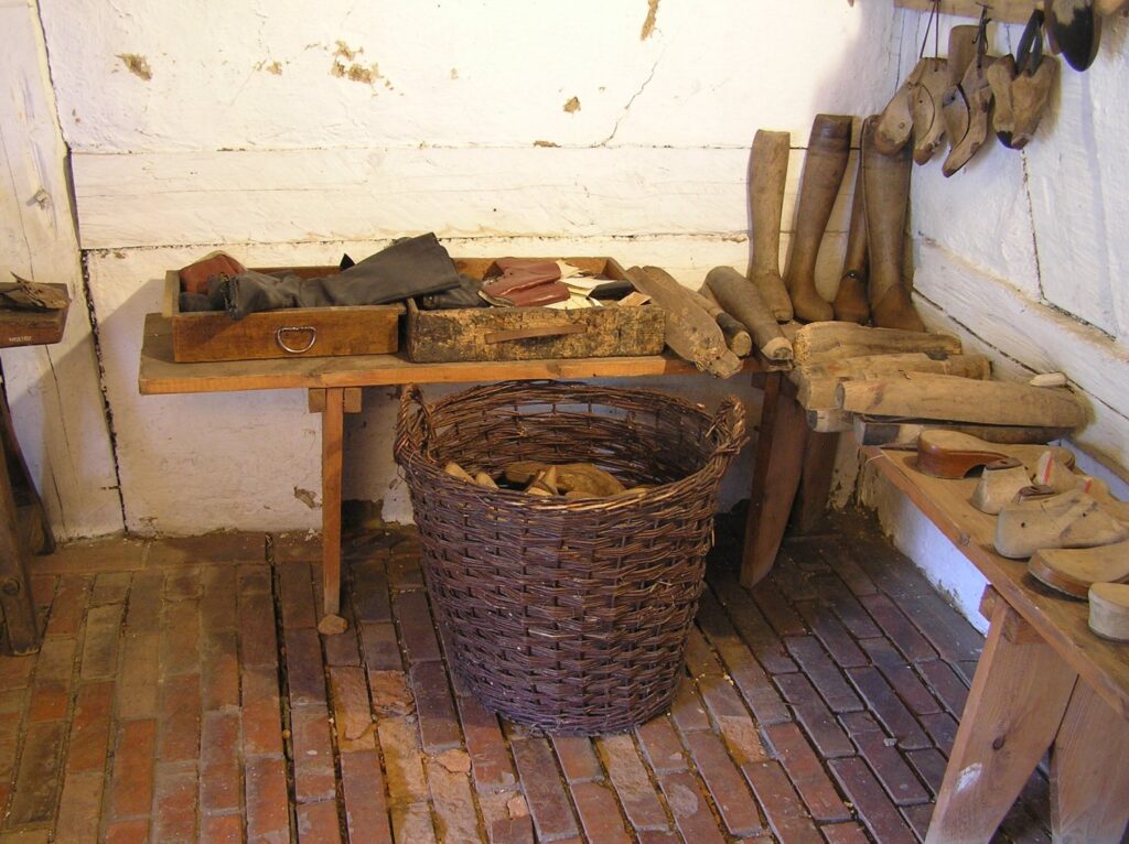 Ława w warsztacie szewskim z narzędziami do obróbki skóry, drewnianymi kopytami oraz koszem z materiałami (kliknięcie powiększy zdjęcie).