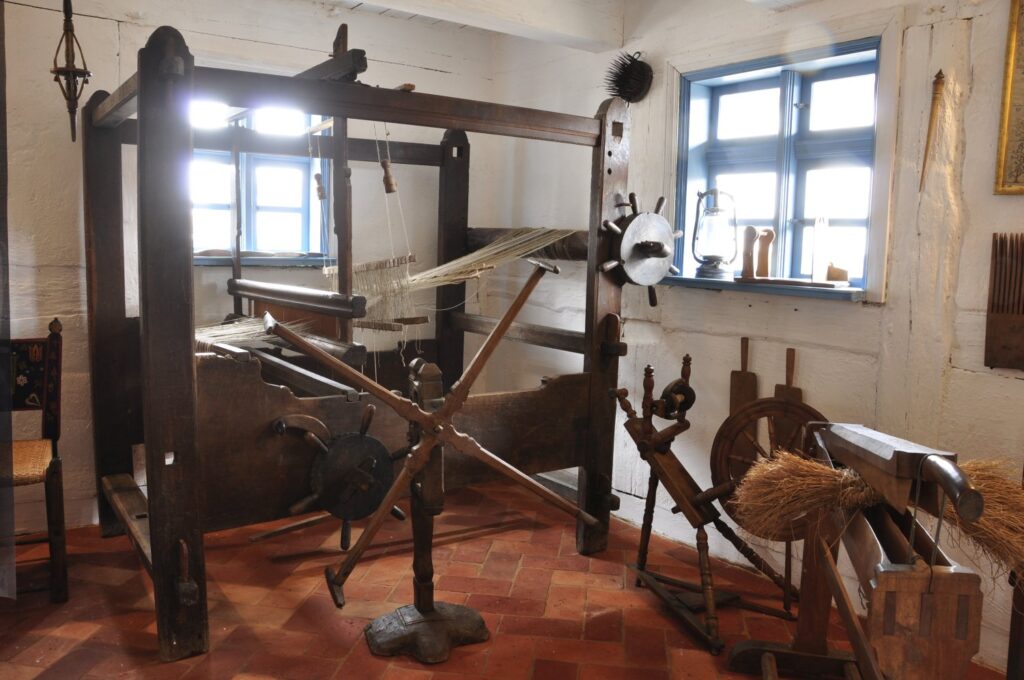 Wnętrze dawnego warsztatu z krosnem tkackim i narzędziami rzemieślniczymi, oświetlone światłem z okna (kliknięcie powiększy zdjęcie).