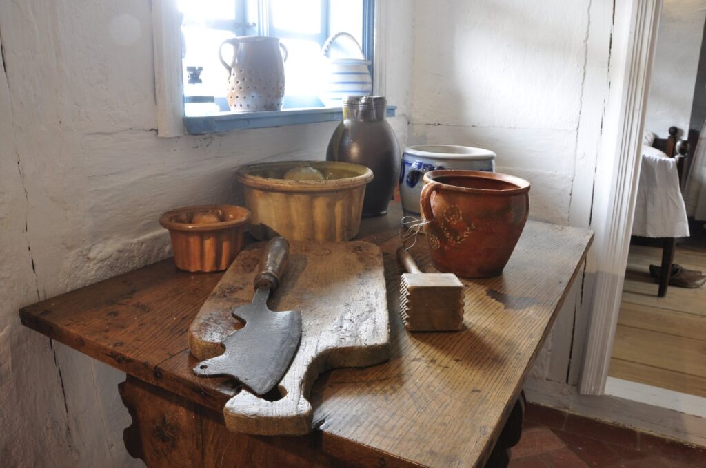 Drewniany stół z deską do krojenia, nożem i glinianymi naczyniami ustawionymi przy oknie w dawnej izbie (kliknięcie powiększy zdjęcie).