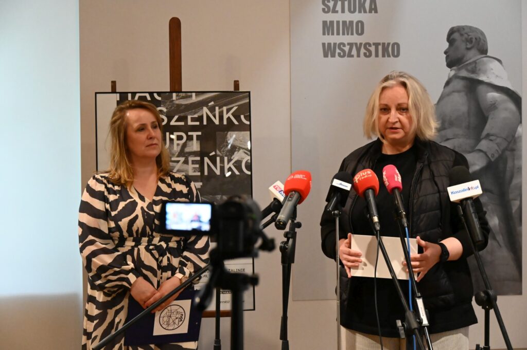 Joanna Chojecka i Katarzyna Radecka na konferencji prasowej poświęconej "Bezpiecznym Wakacjom 2025". W tle plakat wystawy "Sztuka mimo wszystko".