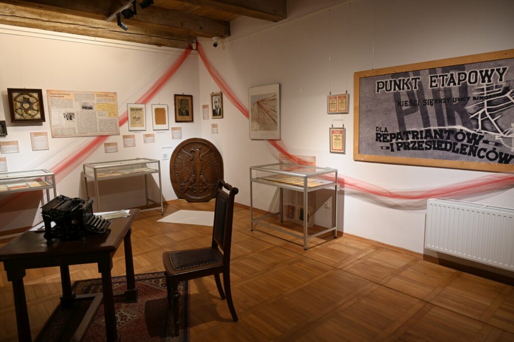 Tablica z napisem "Punkt etapowy dla repatriantów i przesiedleńców". Po lewej stronie dokumenty i tablice opisujące historię miasta na krótko po zakończeniu II wojny światowej.