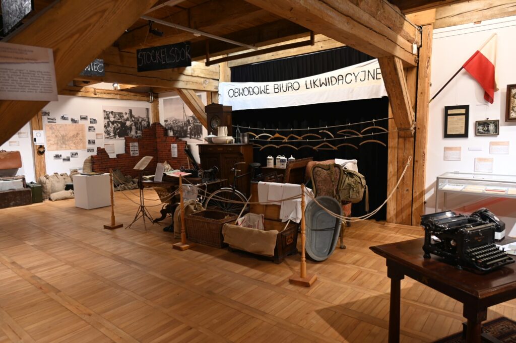 W tle napis "Obwodowe biuro likwidacyjne", a przed nim rzeczy codziennego użytku: rower, meble, porcelana, maszyna do szycia itp.