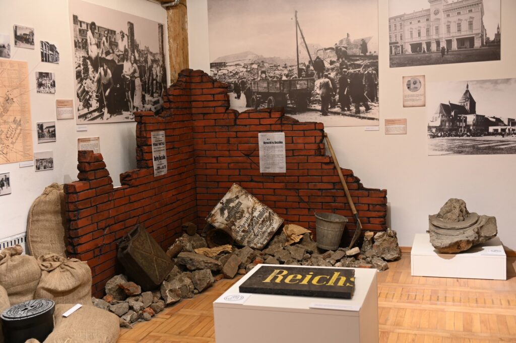 Fragment tablicy z niemieckiego banku Reichsbank, a za nim ceglany mur i wywieszone archiwalne zdjęcia dokumentujące okres odbudowy Koszalina po II wojnie światowej.